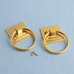 K&ouml;nnte beinhalten: Zwei goldfarbene Ringgriffe mit quadratischen Grundplatten und einer kleinen Schraube auf hellblauem Untergrund. Die Ringgriffe sind wahrscheinlich f&uuml;r M&ouml;bel oder Schr&auml;nke gedacht und bieten ein klassisches Design.