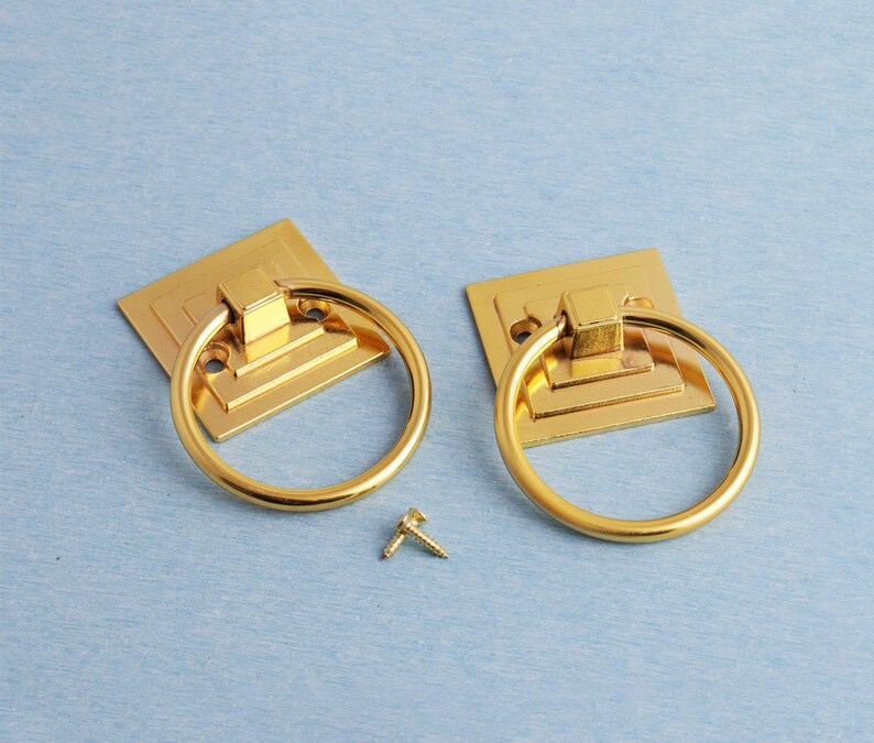 K&ouml;nnte beinhalten: Zwei goldfarbene Ringgriffe mit quadratischen Grundplatten und einer kleinen Schraube auf hellblauem Untergrund. Die Ringgriffe sind wahrscheinlich f&uuml;r M&ouml;bel oder Schr&auml;nke gedacht und bieten ein klassisches Design.