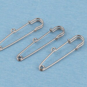 May include: Three silver-toned metal safety pins, each featuring a small circular loop integrated into the side of the pin shaft. These fasteners have a classic coil spring mechanism at the top and a pointed end secured within a protective clasp.