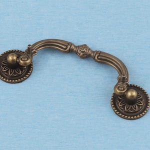 May include: An antique brass-toned metal drawer pull featuring a curved handle with a central decorative element. The handle is attached to two circular backplates, each adorned with a detailed floral pattern and a beaded outer edge border.