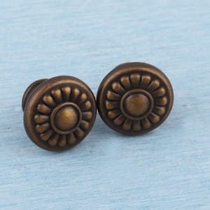 May include: Two antique bronze cabinet knobs with a floral design. Each knob features a central circle surrounded by a ring of petal-like shapes. The knobs are set against a light blue background.