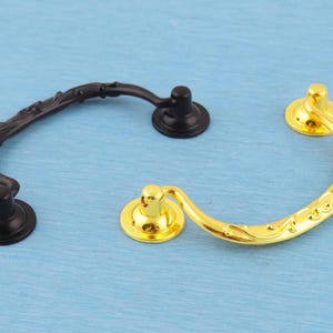 Könnte beinhalten: Zwei Schubladengriffe aus Metall, einer in mattschwarzer Ausführung und einer in polierter Goldoptik. Jeder Griff verfügt über einen geschwungenen, beweglichen Bügel mit geprägtem Blumenmuster und zwei runde Befestigungssockel.