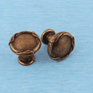 Puede incluir: Dos pomos redondos para armarios con acabado en bronce oscuro y un aspecto texturizado y desgastado. Cada pomo presenta un borde elevado en forma de hoja que rodea un centro liso y ligeramente abombado, con una base cilíndrica corta.