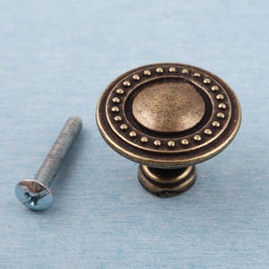 May include: An antique brass cabinet knob with a circular design. It has a central disc and a dotted outer ring. A silver screw is next to the knob. The knob is for furniture and home decor.
