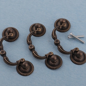 Puede incluir: Tres tiradores de cajón de bronce antiguo con detalles decorativos florales y de volutas. Cada tirador tiene un asa curva y se incluyen dos tornillos de montaje. Los tiradores están colocados sobre un fondo azul claro.