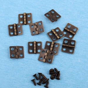 Può includere: Una collezione di piccole cerniere metalliche rettangolari color bronzo antico, ciascuna dotata di quattro fori circolari per le viti. Accompagnano le cerniere un piccolo mucchio di viti metalliche abbinate color bronzo con punte.