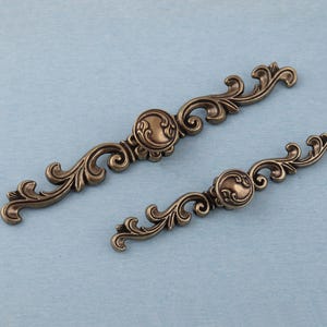 Può includere: Due maniglie per mobili color bronzo antico con disegni a volute ornate. Ogni maniglia presenta un medaglione centrale circolare e bracci curvi e simmetrici. La maniglia più grande misura circa 20 cm di lunghezza, quella più piccola circa 13 cm.