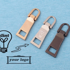 May include: Three metal zipper pulls in rose gold, silver, and dark gunmetal tones. Each pull features a rectangular body with a square cutout at the bottom and a hinged loop at the top. The silver pull is engraved with a lightbulb icon and the text glint.
