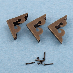 Puede incluir: Tres escuadras de esquina de color bronce antiguo con adornos decorativos, junto con varios clavos pequeños. Las escuadras en forma de L están diseñadas para reforzar las esquinas. El hardware se muestra sobre una superficie azul claro.
