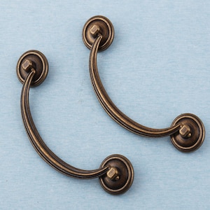 Op de afbeelding: Twee antieke bronzen ladehandgrepen. Elke handgreep heeft een gebogen handvat met een decoratief ontwerp en cirkelvormige uiteinden. Het beslag staat tegen een lichtblauwe achtergrond, wat het vintage-stijl ontwerp benadrukt.