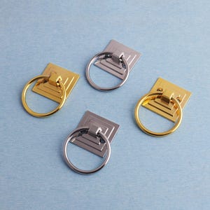 May include: Four cabinet pulls with square backplates and ring handles. Two are gold-toned, and two are silver-toned. The pulls are arranged on a light blue surface, offering a contrast of metallic finishes.