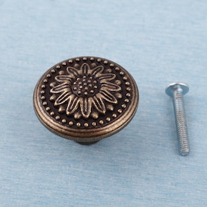 May include: A bronze-colored cabinet knob with a sunflower design. The knob has a round shape with a detailed sunflower in the center, surrounded by a dotted border. A silver screw is next to the knob.