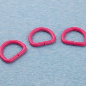 May include: Three bright pink D-rings are arranged on a light blue surface. Each D-ring has a smooth, rounded shape with a small, visible gap on the straight side. The rings are evenly spaced, creating a simple, clean composition.