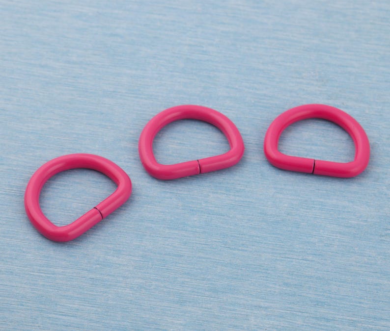 Pu&ograve; includere: Tre anelli a D di colore rosa acceso sono disposti su una superficie azzurra. Ogni anello ha una forma liscia e arrotondata con una piccola fessura visibile sul lato dritto. Gli anelli sono ben distanziati, creando una composizione pulita.