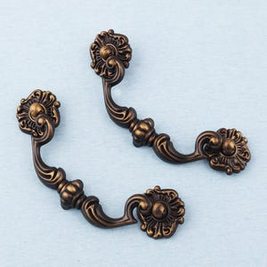 Könnte beinhalten: Zwei verzierte Schubladengriffe in Bronzefarbe mit floralem Design. Jeder Griff hat einen gebogenen Griff und dekorative Endstücke. Die Griffe sind vor einem hellblauen Hintergrund platziert, der ihre komplizierten Details hervorhebt.