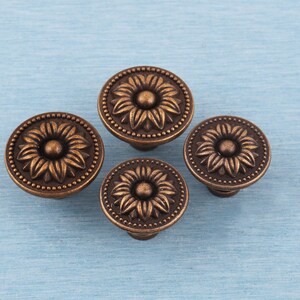 May include: Four bronze cabinet knobs with a sunflower design. Each knob features a central flower surrounded by detailed petals and a dotted border. The knobs are arranged on a light blue surface.
