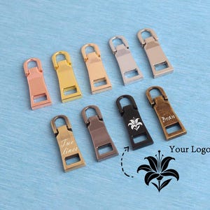 May include: A collection of nine metal zipper pulls in various finishes including rose gold, gold, silver, copper, black, and antique brass. Some pulls feature custom engravings like a lily flower, the name Bran, or the text Two lines.