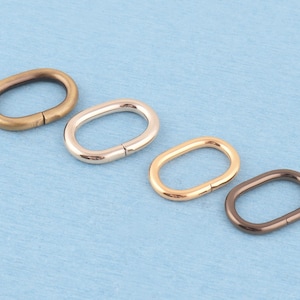 Peut inclure: Quatre anneaux métalliques de forme ovale disposés en ligne diagonale. Chaque anneau présente une finition métallique différente, notamment laiton antique, argent poli, or brillant et bronze à canon, avec une couture visible sur chaque boucle.