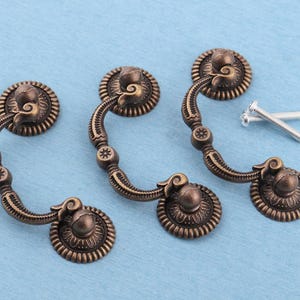 Peut inclure: Trois poignées de tiroir en métal couleur bronze antique avec des anses courbées et des plaques de montage circulaires ornées de motifs d'étoiles et de rayons, accompagnées de deux vis de fixation en métal argenté avec tiges filetées.