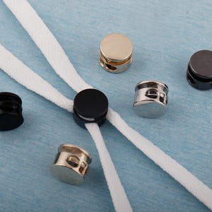 Könnte beinhalten: Eine Auswahl an Kordelstoppern in Gold, Silber und Schwarz, mit weißen Kordeln, die durch sie gefädelt sind. Die Kordelstopper sind zylindrisch mit zwei Löchern für die Kordel. Der Hintergrund ist hellblau.