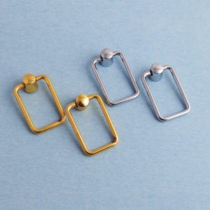 以下が含まれることがあります： 金と銀色の4つのキャビネットプル。各プルは、上部に円形のノブが付いた長方形のフレームを備えています。淡い青色の表面に配置され、デザインと仕上げを際立たせています。