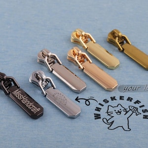 May include: An assortment of metal zipper pulls in black, silver, gold, and rose gold. Some pulls have engraved text, and a logo is visible. The pulls are displayed on a blue surface.
