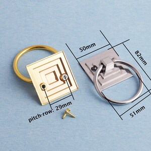 K&ouml;nnte beinhalten: Zwei Schrankgriffe, einer goldfarben und einer silberfarben, mit Ringgriffen. Der goldfarbene Griff hat eine quadratische R&uuml;ckplatte mit geometrischem Design und einem Lochabstand von 29 mm. Der silberfarbene Griff misst 50 mm x 82 mm x 51 mm. Inklusive Schrauben.