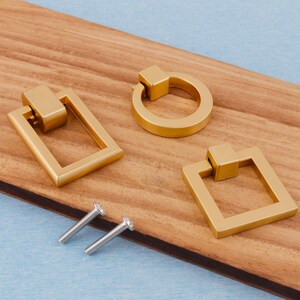 Op de afbeelding: Drie goudkleurige kastgrepen in verschillende vormen: een rechthoek, een cirkel en een vierkant. Er zijn ook twee zilveren schroeven aanwezig. De grepen liggen op een houten oppervlak met een blauwe achtergrond.