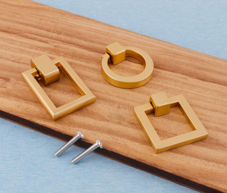 Op de afbeelding: Drie goudkleurige kastgrepen in verschillende vormen: een rechthoek, een cirkel en een vierkant. Er zijn ook twee zilveren schroeven aanwezig. De grepen liggen op een houten oppervlak met een blauwe achtergrond.