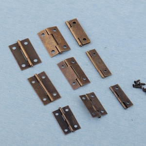 Może przedstawiać: Zestaw zawiasów w kolorze antycznego brązu i małych śrub. Zawiasy są prostokątne z zaokrąglonymi narożnikami i mają dwa skrzydła połączone centralnym sworzniem. Śruby są małe i ciemne.