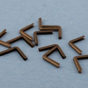 May include: A collection of bronze-colored, right-angle metal corner protectors. These L-shaped pieces are designed to reinforce the corners of various craft or woodworking projects. The protectors are scattered on a light blue surface.