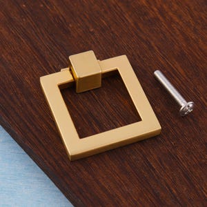 Op de afbeelding: Een goudkleurige vierkante ladeknop met een bijpassend vierkant middenstuk. Een zilveren schroef is naast de ladeknop geplaatst. De hardware is ontworpen voor kasten en laden.