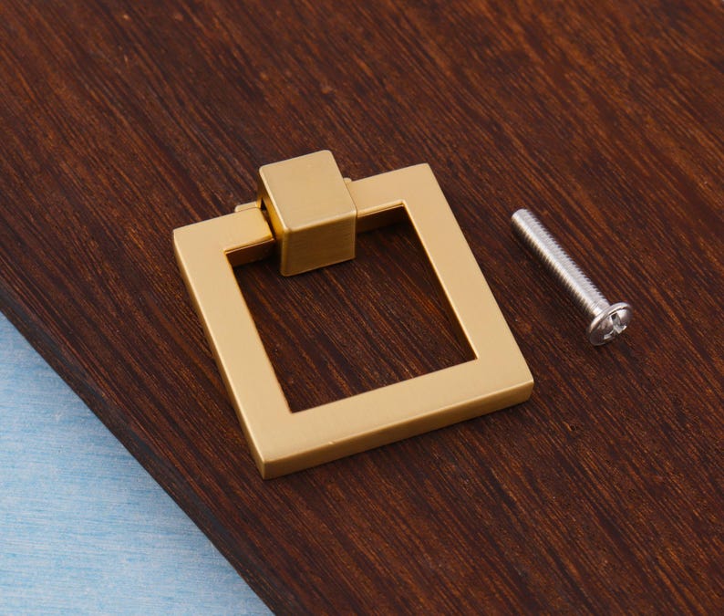 Op de afbeelding: Een goudkleurige vierkante ladeknop met een bijpassend vierkant middenstuk. Een zilveren schroef is naast de ladeknop geplaatst. De hardware is ontworpen voor kasten en laden.