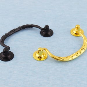 May include: Two decorative drawer pulls. One is black with a curved handle and floral design, the other is gold-coloured with a similar shape and ornate detailing. Both have circular mounting plates.