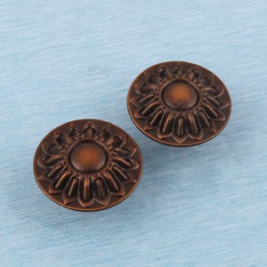 May include: Two bronze-colored cabinet knobs with a sunflower design. Each knob is round with a central circle surrounded by petal-like details. The knobs are set against a light blue, textured background.