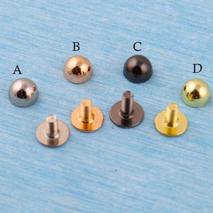 May include: A collection of four metal dome rivets in silver, gold, black, and brass colors. Each rivet has a rounded head and a post for attachment. The rivets are displayed on a light blue surface.