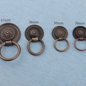 Könnte beinhalten: Vier antike bronzefarbene Schrankgriffe mit Ringgriffen. Die Griffe haben ein kreisförmiges Design mit einem floralen Muster in der Mitte. Die Größen sind 47 mm, 36 mm und zwei mit 30 mm.