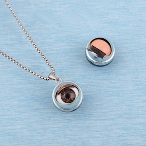 May include: A silver-toned necklace with a pendant featuring a detailed brown eye. Beside it, a small silver container holds a pink object and a black strip. The items rest on a light blue surface.