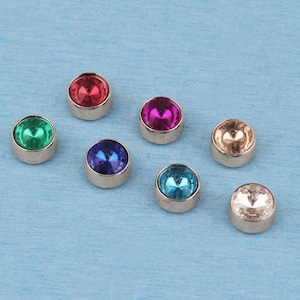 May include: A collection of seven circular metal studs, each featuring a faceted gemstone in a different color. The set includes red, magenta, green, blue, turquoise, champagne, and clear stones, all set within a polished silver-toned metal casing.