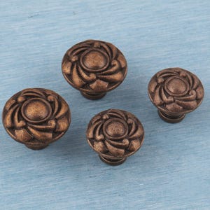 May include: Four bronze-colored cabinet knobs with a floral design. The knobs are round with a raised, swirling pattern and a central circular detail. They are set against a light blue background.