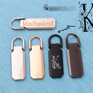 May include: A set of five rectangular metal zip pulls with rounded tops and attached loops. One pull features the text MissHandcraft, while another displays a floral letter K design. The pulls are finished in silver, gold, black, and gunmetal tones.