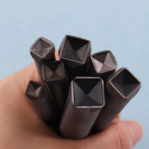 Puede incluir: Un conjunto de herramientas de metal oscuro con puntas cuadradas, posiblemente para trabajos en cuero o estampado de metal. Las herramientas tienen un diseño geométrico en el extremo cuadrado y se sostienen en una mano sobre un fondo azul claro.