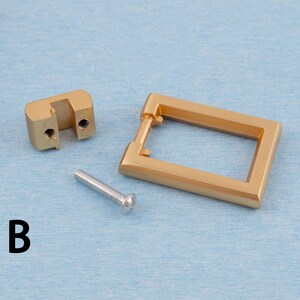 Op de afbeelding: Een set goudkleurige kastbeslagonderdelen, waaronder een rechthoekige trekker, een montageblok en een zilveren schroef. De hardware wordt weergegeven op een lichtblauwe ondergrond. De letter "B" is in het zwart gedrukt.