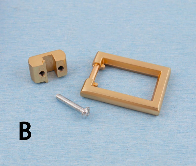 Op de afbeelding: Een set goudkleurige kastbeslagonderdelen, waaronder een rechthoekige trekker, een montageblok en een zilveren schroef. De hardware wordt weergegeven op een lichtblauwe ondergrond. De letter "B" is in het zwart gedrukt.