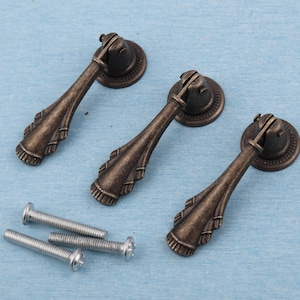 May include: Three antique bronze drawer pulls with decorative detailing and matching screws. The pulls have a classic drop handle design and are set against a light blue background. Ideal for furniture restoration projects.