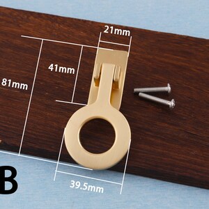 Puede incluir: Un tirador de armario dorado con un asa en forma de anillo, de 39,5 mm de ancho y 81 mm de largo. Incluye dos tornillos plateados para la instalaci&oacute;n. Montado sobre una superficie de madera marr&oacute;n oscuro.
