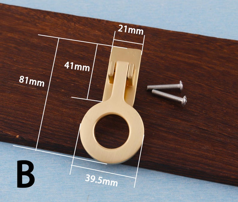 Puede incluir: Un tirador de armario dorado con un asa en forma de anillo, de 39,5 mm de ancho y 81 mm de largo. Incluye dos tornillos plateados para la instalaci&oacute;n. Montado sobre una superficie de madera marr&oacute;n oscuro.