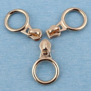 Puede incluir: Tres tiradores de cremallera de color dorado con un diseño circular. Los tiradores están conectados entre sí.