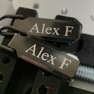 K&ouml;nnte beinhalten: Zwei schwarze Metallanh&auml;nger mit der Gravur "Alex F". Die Anh&auml;nger haben eine rechteckige Form mit abgerundeten Kanten und einer kleinen Schlaufe. Der Text ist in einer klaren, serifenlosen Schriftart.