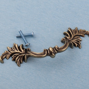 May include: An antique-style bronze drawer pull with ornate leaf detailing. Includes two silver screws for installation. The curved handle is approximately 13 cm long, suitable for cabinets and furniture restoration projects.